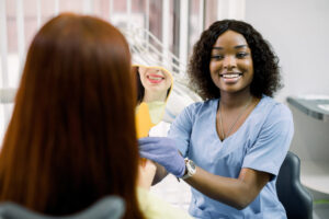 Back view of woman patient looking at her smile after curing or whitening teeth in dental clinic, and happy African female dentist, showing the mirror with reflection of smile