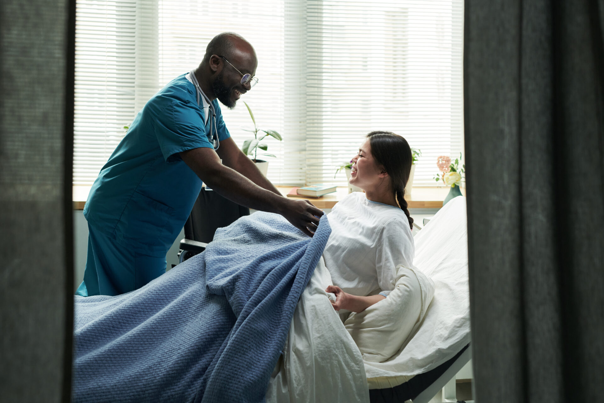 Black Male Nurse Covering Pregnant Caucasian Woman with Blanket in Ward