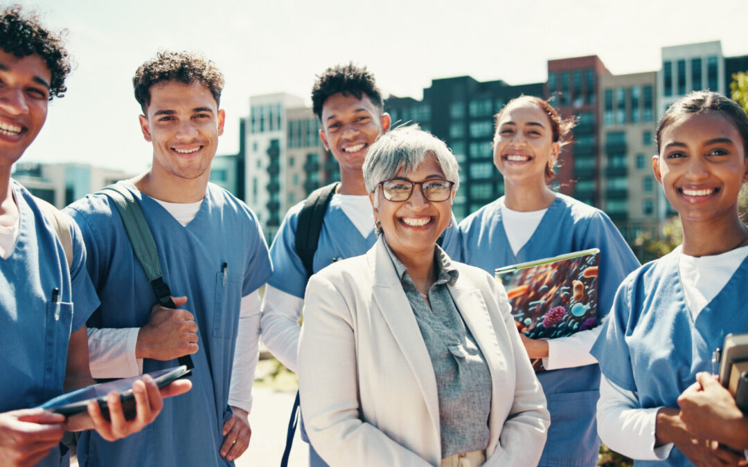 A group of 5 medical students stand outside a university smiling with their professor who stand in front of them in a white lab jacket.