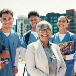 A group of 5 medical students stand outside a university smiling with their professor who stand in front of them in a white lab jacket.