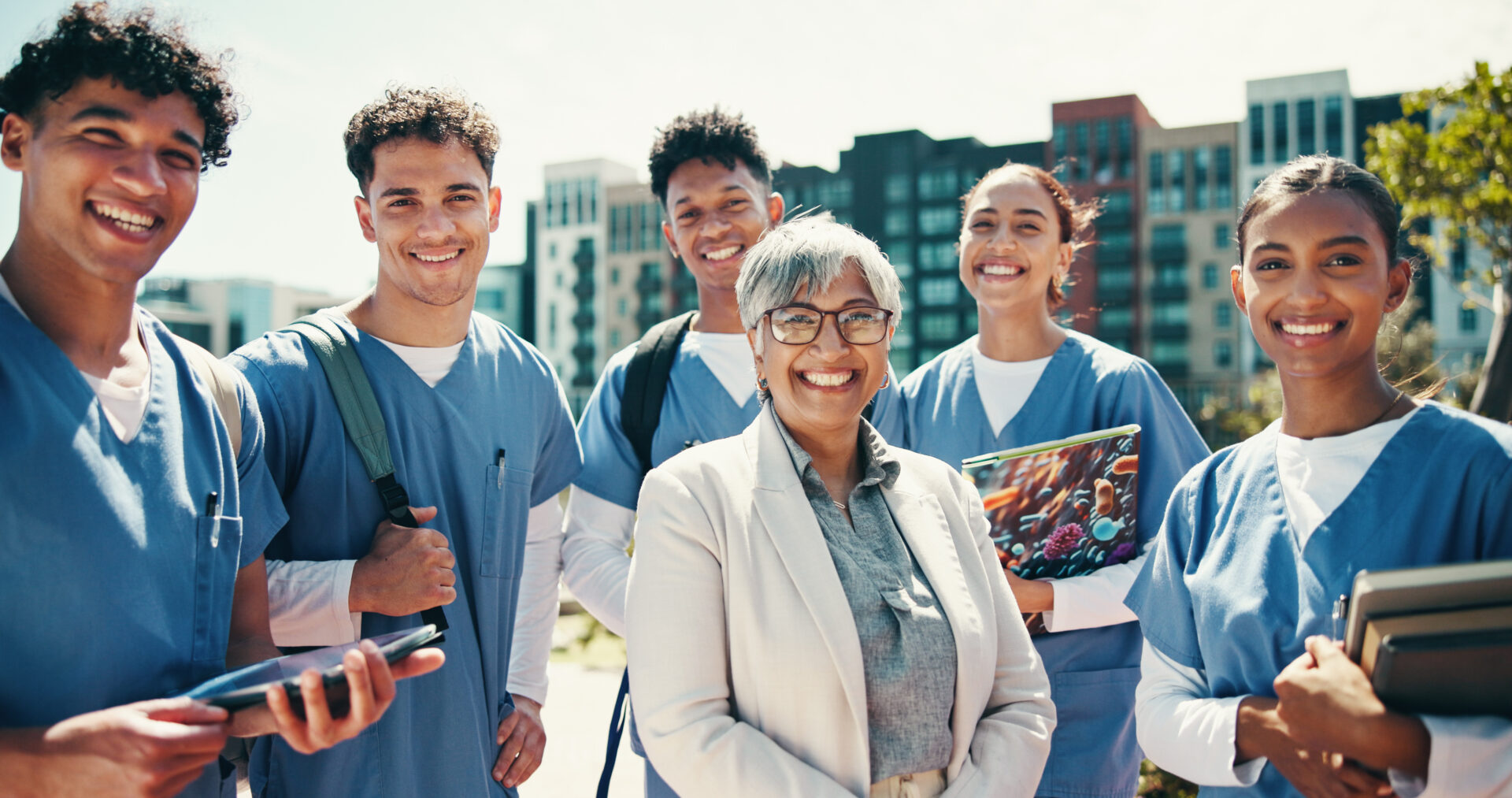 smiling-medical-professionals-standing-outside-bui-2026-03-25-02-15-32-utc A group of 5 medical students stand outside a university smiling with their professor who stand in front of them in a white lab jacket.